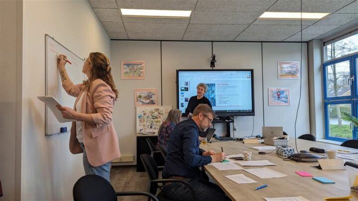 Kantoorruimte waarin een vrouw op whiteboard schrijft. Andere personen bestuderen documenten aan tafel.