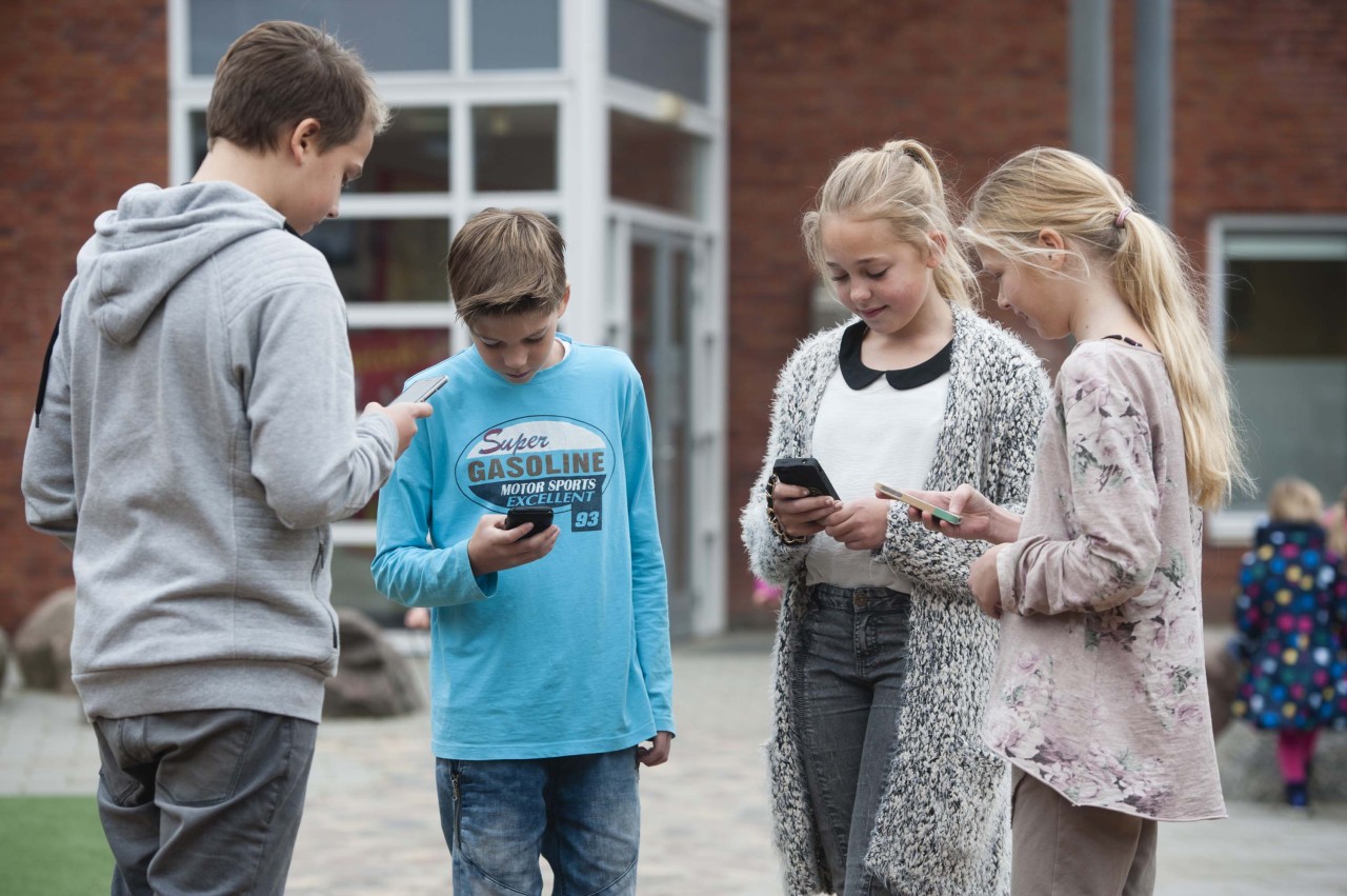 Een groepje kinderen staat op het schoolplein, ze kijken allemaal naar hun eigen mobiele telefoon