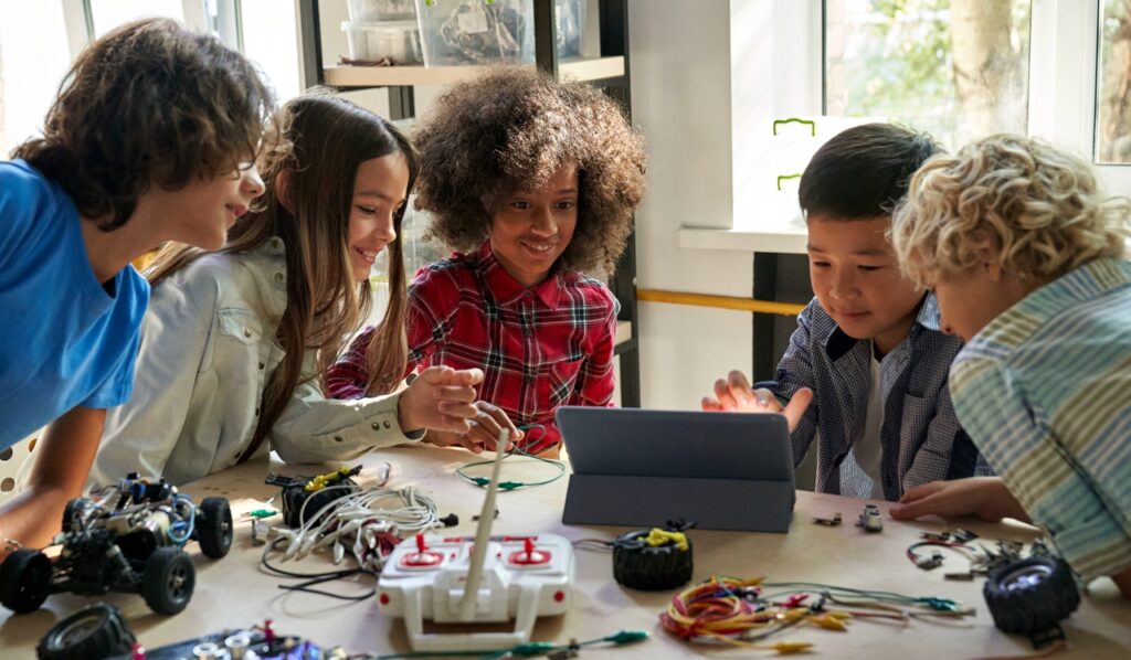 Vijf vrolijke kinderen rondom tafel met tablet, kabeltjes, joystick en robotautootje